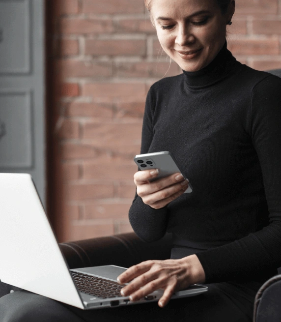 Smiling woman working on a laptop with a smartphone for transcription services to handle diverse speech patterns and accents. Smiling woman working on a laptop with a smartphone for transcription services to handle diverse speech patterns and accents.